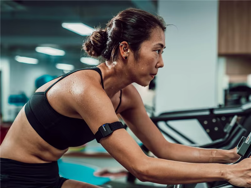 A cyclist gripping handlebars, with the COOSPO HW9 visible on their bicep, the LED glowing green for moderate intensity.