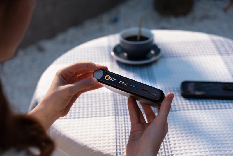 A photographer's hand inserting an SD card into the side of the PocketCloud device, which is resting on a rock or outdoor table, with a camera blurred in the background.
