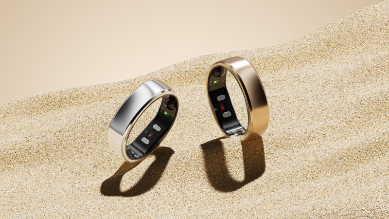 A detailed product photograph of two RingConn Gen 2 rings resting side-by-side on a textured neutral surface. One ring is a brushed silver finish, and the other is a polished gold finish, showing the unique "squircle" shape.